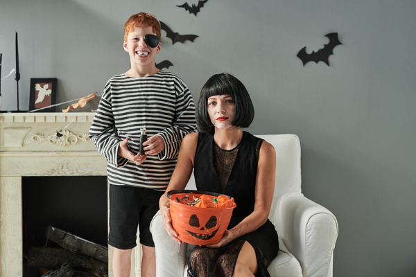 Boy in Pirate Costume Stands next to His Mother in Witch Costume Which Holds Bucket of Halloween Treats